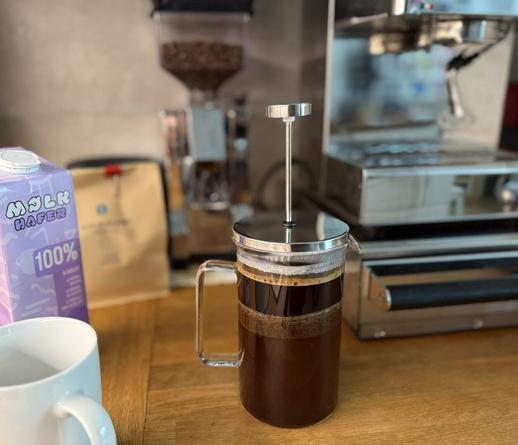 Eine mit gebrühtem Kaffee gefüllte French Press steht auf einer Holzfläche, im Hintergrund befinden sich eine Kaffeemühle & eine Espressomaschine. An der Seite steht eine Packung Hafermilch neben einem weißen Kaffee-Becher