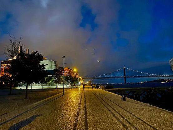 A scenic nighttime view of a waterfront promenade featuring a bridge illuminated by lights. Nearby buildings, partially obscured by trees, are softly lit, and a pathway lined with cobblestones leads toward the water. The sky is cloudy with hints of color,