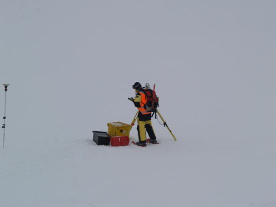 A person in a hi viz vest and yellow/black clothing carrying a backpack and looking at a handheld instrument. The person stands in front of a surveying tripod holding a robotic total station. A few boxes are in front of the person, a red box for the total station, yellow box holding a large battery and warming equipment (a hot water bottle), custom made by the person in the photo. The black box holds extra equipment that isn't cold sensitive. On the far left a pole with a GNSS antenna and total station prism is stuck into snow. The foreground and background are white - a foggy day on snow, in the East Antarctic pack ice zone. Photo by Dr. Jan Lieser, subject is Dr. Adam Steer.