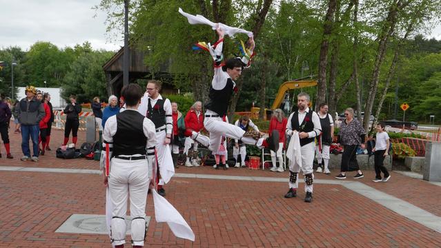 Four morris dancers with white shirts and trousers and black vests, decorated with multicolor ribbons, dancing on a brick paved surface. Three are standing and one is leaping high with arms raised. Behind them an audience including some other morris dancers.