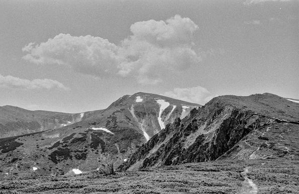 This is a black and white film photograph taken in the Carpathian Mountains, specifically along the Chornohora Ridge. The image features a dramatic mountain landscape with rugged rocky slopes in the foreground and a prominent peak in the distance, partially covered with patches of snow. Puffy clouds drift across the sky above the mountains, adding texture to the scene. The film grain is clearly visible, giving the photo a classic, nostalgic feel and emphasizing its analog origin. The path in the lower right corner suggests a hiking trail, drawing the viewer’s eye into the mountainous terrain.