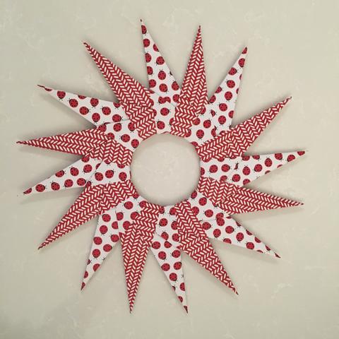 Radial paper wreath with alternating red chevron and ladybug patterns on pointed segments, creating a vibrant, playful design on a light background.
