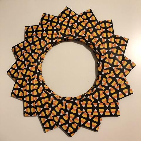 A circular wreath made of interwoven paper with a candy corn pattern. The vivid orange, yellow, and white colors rest against a black backdrop.