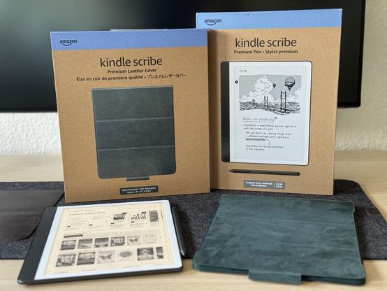 Kindle Scribe 2024 mit passender smaragdgrüner Lederhülle und entsprechenden Verpackungen liegen auf einem Schreibtisch vor einem ausgeschalteten Monitor.