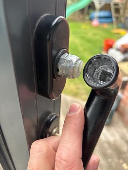 A black door handle that has come off of the piece that attaches it to the spindle. The spindle attachment is protruding from a vertically mounted pill-shaped (straight sides, rounded top and bottom) escutcheon with a separate escutcheon for the key just visible lower down.