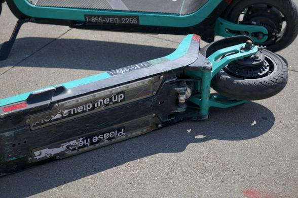Looking down at a rental scooter on its side. The stickers on the bottom read “Please help me up” but are ripped so they’re hard to read. An upright scooter is in the background.