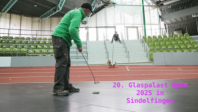 Screenshot aus dem Video zum Crossgolf Turnier in Sindelfingen. Darauf steht ein Mann in einer Turnhalle auch dem mit Platten belegten Hallenboden und puttet einen Golfball in Richtung eines Kinder Hüpftiers