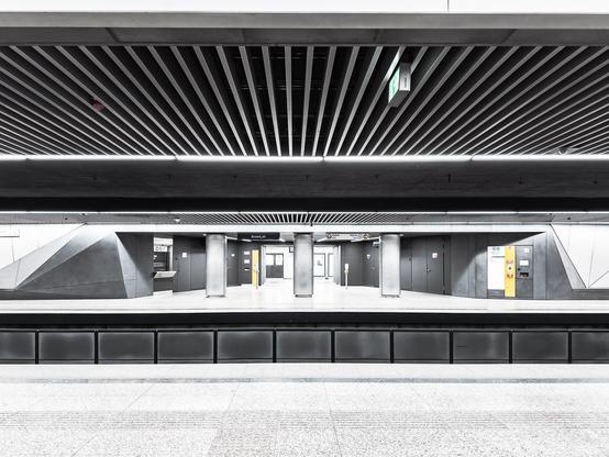 Die Aufnahme zeigt eine moderne, saubere und gut beleuchtete U-Bahn-Station. Die Architektur ist schlicht und zeitgemäß, mit grauen und schwarzen Tönen sowie einer Decke mit parallelen Lamellen und heller Beleuchtung. Strukturelle Säulen und Hinweisschilder zur Orientierung sind sichtbar. Die Plattform ist leer, es sind keine Menschen zu sehen. Der Gesamteindruck ist minimalistisch und offen, wodurch die räumliche Weite und das Design der Station betont werden.