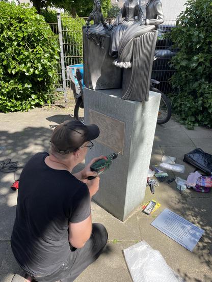 a man with a basecap (me) kneels in front of a granite block and drills a hole in the stone with a percussion machine. A sculpture made of aluminium is recognisable on the block: three female figures sit on the upper edge of an open book