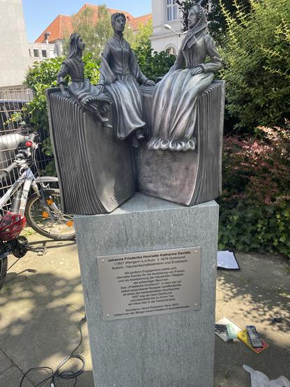 The monument in honour of Henriette Davidis, the author of the first German-language cookbook. There is an aluminium sculpture on the granite plinth with the text plaque. It shows an open book standing upright. Three female figures of different ages sit on the upper edge of the book. They represent Henriette Davidis as a teenager, an adult and an old woman
