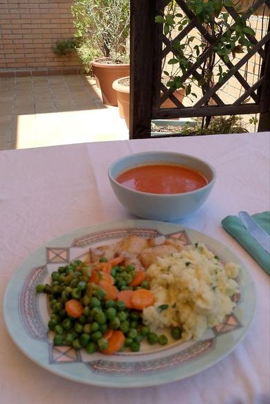 Una mesa rectangular con un mantel blanco en una terraza. Se ven algunos macetones con plantas y una celosía de madera en la que se enreda un jazmín chino.
Sobre la mesa hay un tazón azul claro con gazpacho y un plato plano con dos filetes de pollo, guisantes, zanahorias y un puré de patatas con perejil.