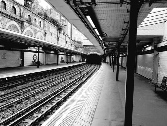 A black and white photo of the Sloan Square tube station. No trains, just empty tracks. No people save one gentleman seated on the opposite side.