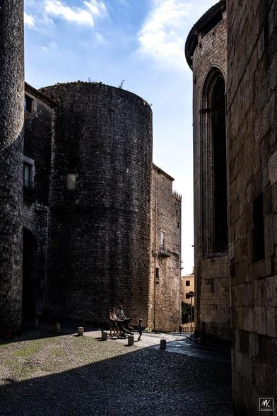 Color photo of a couple walking with each pushing a stroller with a child inside, and a third child walking in front of them, through a patch of sunlight with darkly shadowed old stone buildings on either side.