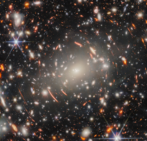 From source in toot: A field of galaxies in space, dominated by an enormous, bright-white elliptical galaxy that is the core of a massive galaxy cluster. Many other elliptical galaxies can be seen around it. Also around it are short, curved, glowing red lines, which are images of distant background galaxies magnified and warped by gravitational lensing. A couple of foreground stars appear large and bright with long spikes around them