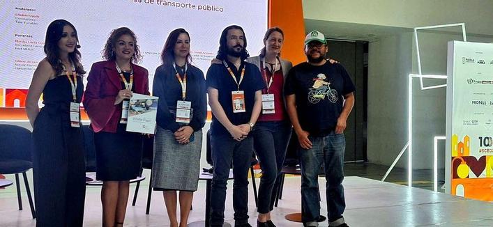 Six panelists standing together at a transportation conference, wearing conference badges and lanyards. From left to right: a woman in a black dress, a woman in a burgundy blazer holding what appears to be an award or certificate, a woman in a dark blue blazer, a bearded man in a dark blue shirt, a woman in a red blazer, and a man in a black t-shirt with a bicycle graphic and baseball cap (Juan Manuel Berdeja Maldonado). Behind them is a presentation screen with Spanish text about public transportation, and conference signage is visible in the background.