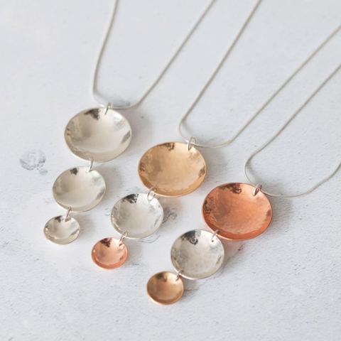 Three necklaces laid out side-by-side on a white surface, each made of three domed metal discs of decreasing sizes. One is all silver; the second is brass, silver and copper; the third is copper, silver and brass.