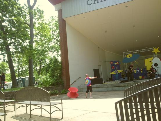 A slightly high-angle, full shot shows a man in a tie-dye shirt standing barefoot on a paved outdoor stage area, facing right with his arm outstretched. Behind him to the right, a band plays on an elevated stage adorned with colorful, whimsical props like a blue UFO and a yellow star. To the left of the man and extending into the background are several tall trees and a row of portable toilets. In the foreground, two empty metal benches are visible on the left, and a dark metal railing leads up to the right. The sun casts strong shadows, suggesting bright daylight.