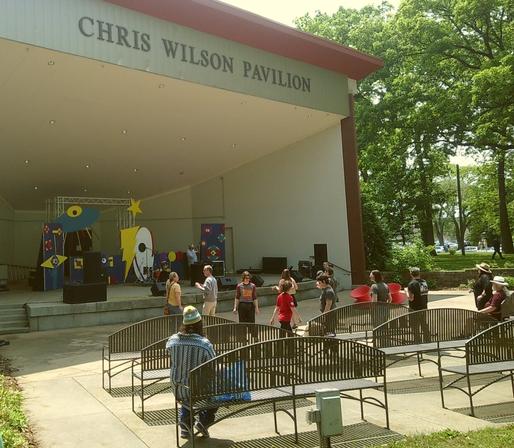A full, eye-level shot shows an outdoor stage area under a covered pavilion with "CHRIS WILSON PAVILION" written across the top. On the stage, whimsical, colorful props resembling a blue UFO, a yellow star, and abstract shapes are set up, and some band equipment is visible.
In front of the stage, on a paved area, several individuals are gathered dancing to the music. One person in the foreground, with their back to the viewer, is seated on a metal bench. There are multiple rows of empty metal benches extending into the middle ground. To the right, a grassy area with many tall trees is visible, and in the far background, a road with a parked car and a person walking can be seen. The scene is brightly lit by the sun, casting shadows from the trees.