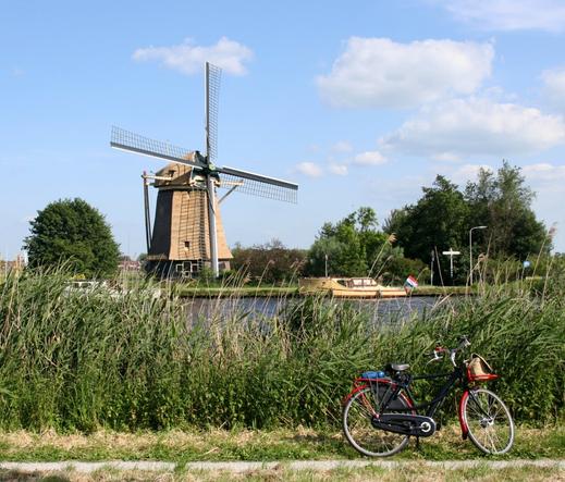 Bike standing alongside of a canal in the countryside. On the other side of the canal there is a brown windmill and a small brown boat with a dutch flag.