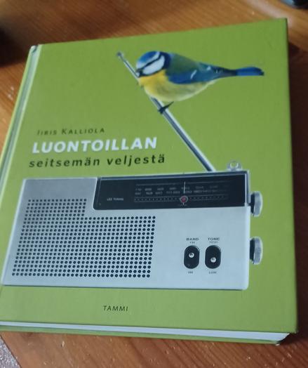 Kuvassa on Iiris Kantolan kirjoittama Luontoillan seitsemän veljestä  kirja puu pöydällä, kirjan kannessa on sinitiainen matkaradion antenilla.