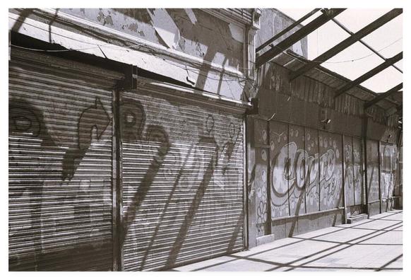 Black and white film photograph showing graffiti on the shuttered properties of an abandoned shopping arcade.