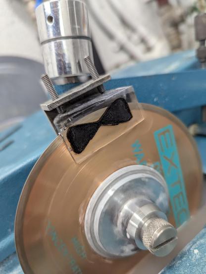 A bow tie shaped piece of dark bone (fossil) embedded in resin being cut by a copper coloured saw blade.