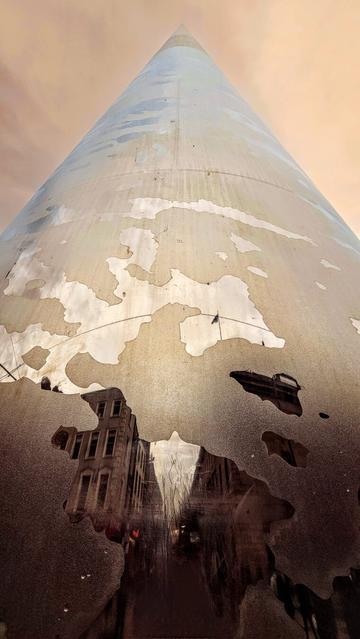 A low-angle shot of the Dublin Spire, a tall, needle-like monument, reflecting the surrounding city and sky. The lower portion of the Spire is visible, showing a textured, weathered surface with patches of rust-like discolouration. Below, the ground is covered in puddles which perfectly mirror the facades of old, Georgian-style buildings and the warm, cloudy sky, creating an intriguing juxtaposition between the monument and its environment. The reflection shows intricate details of the buildings and the subtle colours of the sky, blending seamlessly with the Spire's surface.