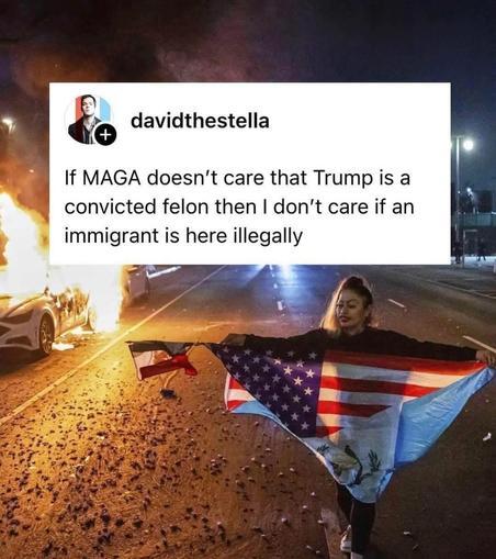 The image shows a person standing on a street at night, holding a large flag that combines elements of the American flag and another flag with a light blue field and a white triangle. The person is wearing a black top and dark pants. In the background, there is a car on fire, with flames and smoke visible. The street is wet, and there are scattered debris and small objects on the ground. The scene is illuminated by streetlights, casting a warm glow over the scene. Overlaid on the image is a text box with a profile picture and the username "davidthetesta," followed by a statement: "If MAGA doesn't care that Trump is a convicted felon then I don't care if an immigrant is here illegally."