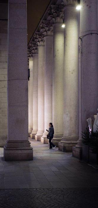 A woman walks among several tall, round columns of a classic-looking building. It's night, and she's illuminated by some lights overhead, while the rest of the scene is darker.