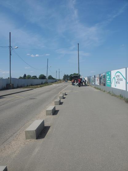 Photo d'une longue route un peu poussiéreuse en plein soleil. La route est entourée de lampadaires et bordée des 2 côtés par des clôtures en tôle laissant penser qu'il y a des travaux. Sur la clôture de droite, on voit des panneaux vantant l'arrivée prochaine du métro. Un peu plus loin, on voit un groupe de personnes patientant à un arrêt de bus