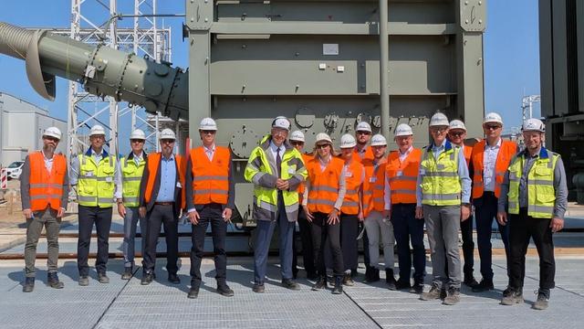 Gruppenbild mit 16 Vertreterinnen und Vertretern von Bundesnetzagentur und Amprion vor dem Konverter in Meerbusch. Die Personen tragen Schutzhelme und Signalwesten.
