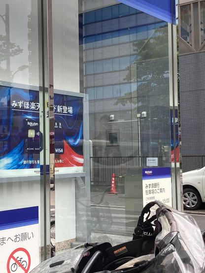 A street scene featuring a glass-encased bank entrance with promotional signage for Mizuho and Rakuten. A stroller is positioned in the foreground, and a traffic cone is visible in the background, along with several buildings reflecting in the glass.