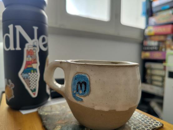 una taza de cerámica con el logo de mastodon junto a un termito con un pegotín de palestina