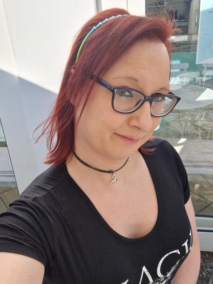 Das Bild zeigt eine Frau mit roten Haaren, die ein schwarzes T-Shirt mit einem weißen Aufdruck trägt. Sie hat eine schwarze Choker-Halskette mit einem silbernen Anhänger um den Hals. Ihre Haare sind mit einem blau-grünen Haarband zusammengehalten. Sie trägt eine Brille mit dunklen Rahmen und lächelt leicht in die Kamera. Im Hintergrund ist ein Glasfenster zu sehen, durch das Sonnenlicht hereinfällt, und ein Teil eines modernen Gebäudes ist sichtbar.