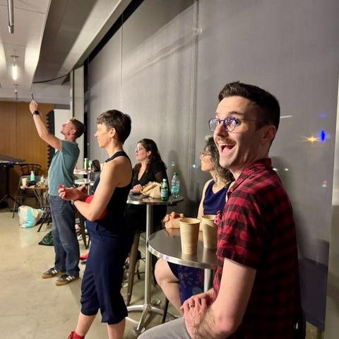 A guy in a red plaid shirt gives the camera a big smile. Several people behind watch the dance floor.
