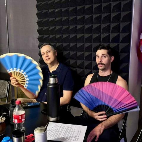 Two guys sit at a table with mate and other drinks in front of them, showing off a pair of large hand fans. One fan is blue with a cream edge. The other has a bi flag design.