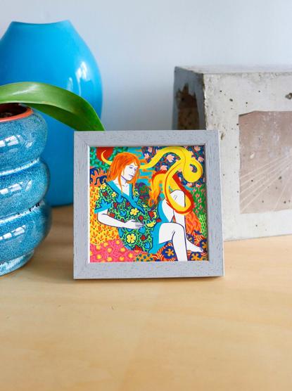 This is a photo of a colored pencil drawing, shown here framed in a small light grey painted wooden frame, placed on a light raw wood surface. On the left, there's a textured blue pot with a plant, and on the right, a carved concrete block. The illustration depicts a woman with bright orange hair, seated in a lush setting. She wears a dress with brightly colored floral patterns and rests her hand on her pregnant belly. A yellow imaginary creature, resembling a small fox with an elongated body, leans in to sniff the woman's belly, much like a companion animal might do. The background is saturated with vegetal and floral patterns in vivid shades of red, green, blue, and orange, creating a dense composition. This image is part of a series of small-format framed illustrations.