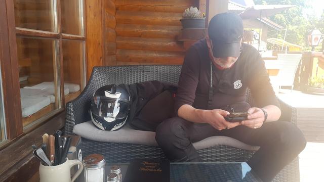 Ein Mann in Motorradklamotten schaut etwas auf dem Smartphone nach. Er sitzt mir gegenüber an einem Couchtisch auf einer Außenterrasse eines Cafés, neben ihm steht ein schwarz-weißer Helm auf der Couch