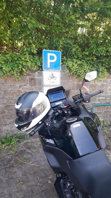 Ein Motorrad steht vor dem Schild mit dem Piktogramm für einen Motorradparkplatz. Am linken Spiegel hängt ein Helm