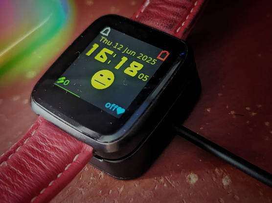 A Pine-Time watch with a red strap in it's charging bay, showing zero battery at 16:18 and a smiley face which isn't smiling.