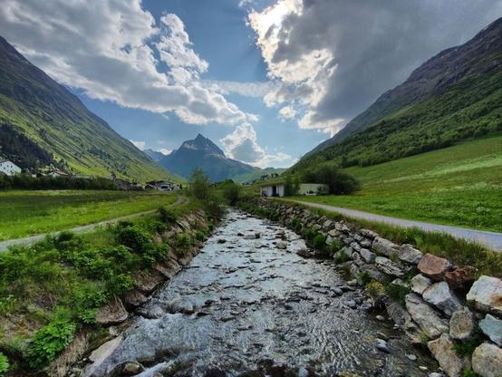 In the foreground, the Vermuntbach flows through Galtür with clear, bubbling water over smooth, moss-covered stones. There are field roads following the river on both sides. In the background, the majestic Ballunspitze rises, its rugged cliffs and green slopes creating a striking contrast to the gentle movement of the water. Snow can be seen in the upper part of the mountain. The sky is a clear blue, dotted with a few grey clouds.