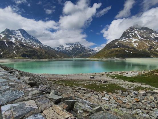 The image captures the stunning Stausee Kops, an alpine reservoir renowned for its striking turquoise hue. On the right hand side a part of its retaining wall can be seen. The lake is surrounded by majestic mountains, some of which are partially blanketed in snow. The clear blue sky is adorned with scattered white clouds, enhancing the vibrant colors of the landscape. In the foreground, a rocky shoreline frames the lake. A tire and some wood debris can be seen along the shore, adding a touch of rustic charm to this picturesque setting.