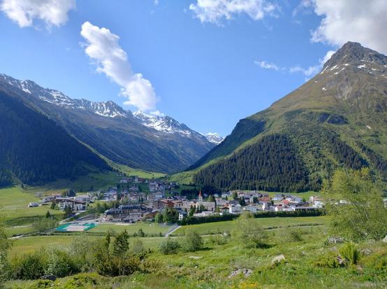 Picturesque view of Galtür nestled in a lush green valley. Surrounded by towering mountains, some capped with snow, the village features various buildings with colorful rooftops. A prominent church with a red steeple stands out among the structures. The sky is bright blue with a few fluffy clouds.