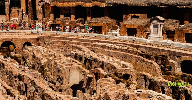 Detailed interior view of the Colosseum in Rome: foreground shows sunlit, ruined hypogeum brick walls and arches; middle ground has a curved marble-edged walkway with tourists; background reveals dark brick arches and a small white monument set against weathered stone.