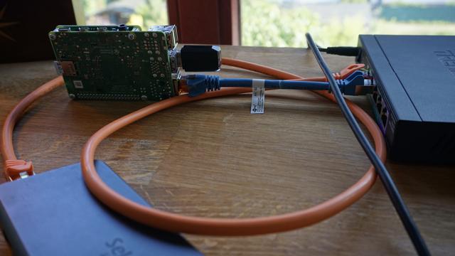 An image of my networking setup. All the gear lays on my wooden desk. The Raspberry Pi is turned on its side. A USB-Wifi-Adapter is connected to it (offering an access point). It is powered over Ethernet. The HAT for PoE+ is installed in a way it is barely visible on the image. The Pi is connected to a Switch, which is connected to power. It is a 5 Port 2.5GbE PoE+ switch. The cable connecting the Pi is blue. Another cable is connected to the switch and a USB-Dock. This cable is orange and longer.