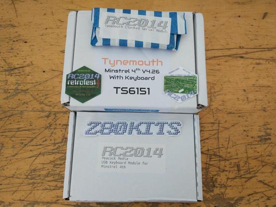 A white cardboard box on a wooden workbench with "Tynemouth Minstrel 4th V4.26 with Keyboard" printed on thew box. A smaller, blue and white striped package rests on top of the box. A label on it reads "Tynemouth Serial Module"

Another, flatter box is placed below them with the text "USB Keyboard Module" prited on it.