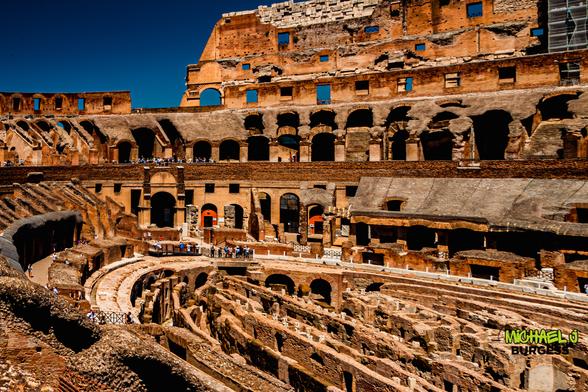 Wide interior view of Rome’s Colosseum showing multiple levels of weathered brick arches, bright blue sky overhead, exposed hypogeum passageways in the foreground, and tourists walking along the mid-level galleries.