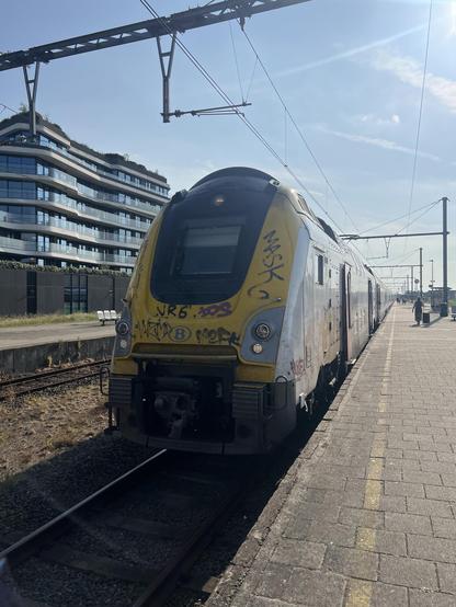 Ein Zug mit Graffiti ist auf den Gleisen neben einem Bahnsteig geparkt, mit einem modernen Gebäude im Hintergrund unter einem klaren blauen Himmel zu sehen.