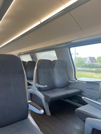 Innenraum eines Zugwagens mit Reihen grau gepolsterter Sitze mit Armlehnen, großen Fenstern und moderner Beleuchtung. Die Aussicht von außen zeigt Grün und ein Wohngebiet.