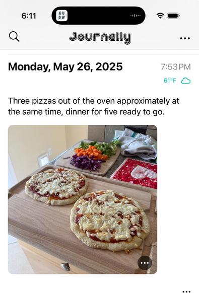 This is a screenshot from an app called Journelly, showing an entry dated Monday, May 26, 2025, at 7:53 PM with a temperature of 61°F. The entry reads: “Three pizzas out of the oven approximately at the same time, dinner for five ready to go.” Below the text, there is a photo showing two homemade pizzas with melted cheese on top sitting on a large wooden cutting board. In the background, there is a kitchen counter with a cutting board holding chopped vegetables, including green leaves, orange and yellow bell peppers, and purple cabbage. There are also folded kitchen towels and a red oven mitt on the counter. The scene suggests a fresh, home-cooked dinner is ready to be served.
